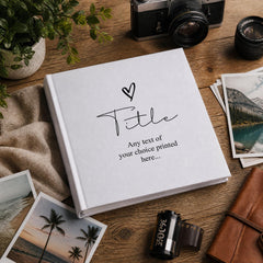 a white photo album with a handwritten title, surrounded by various photography equipment and printed photographs on a wooden surface.