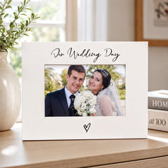 A white picture frame with a photo of a couple, presumably a bride and groom, inside it. The frame has a heart symbol at the bottom. The background of the image is blurred, but it appears to be a room with a window and some books.