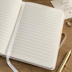 An open notebook with blank lined pages, a pen, and a wedding invitation card are arranged on a wooden surface.