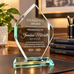 A clear glass award trophy with an inscription, placed on a desk with books and a plant in the background.